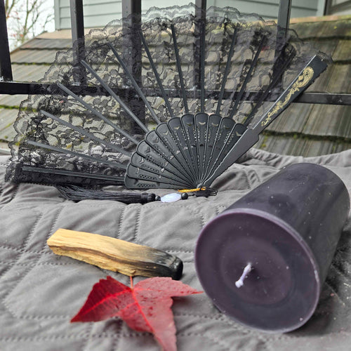 Black lace fan, purple candle, wooden stick, and red leaf on a textured surface with a blurred background.